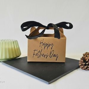 May include: A brown gift box with a black ribbon and the text "Happy Fathers Day". A green candle in a ribbed glass holder and pine cones are on a black square surface.