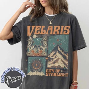 Peut inclure: T-shirt gris foncé avec un motif graphique orange « VELARIS », des illustrations de montagnes, d'étoiles et d'une créature ressemblant à une chauve-souris. Le t-shirt porte également l'inscription « CITY OF STARLIGHT ».