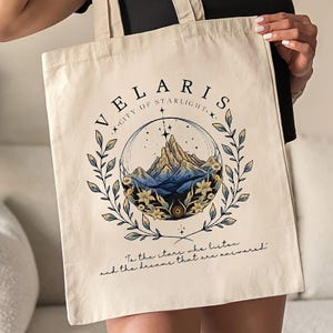 Peut inclure: Sac fourre-tout crème avec un motif de paysage de montagne dans un cercle. Le motif comprend le texte "VELARIS CITY OF STARLIGHT" et la citation "To the stars who listen and the dreams that are answered."