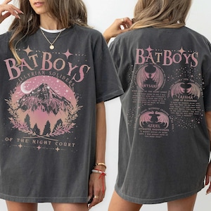 Peut inclure: T-shirt gris foncé avec l'inscription "BATBOYS" en rose, orné d'un motif de montagne et de lune. Le dos du t-shirt porte également l'inscription "BATBOYS" et des illustrations d'ailes et de texte. Le t-shirt est à col rond.