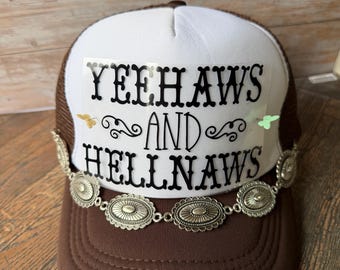Może przedstawiać: Brązowo-biała czapka typu trucker z napisem "YEEHAWS AND HELLNAWS". Czapka posiada ozdobny srebrny łańcuszek z owalnymi zdobieniami i brązowy daszek.