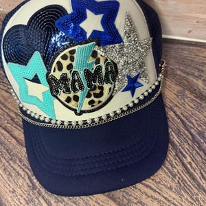 Può includere: Cappellino da camionista blu navy con pannello frontale bianco e visiera curva. Il cappello è impreziosito da stelle di paillettes e strass, un fulmine e la scritta "MAMA" in un motivo leopardato. Un bordo di perle e catena accentua la visiera.