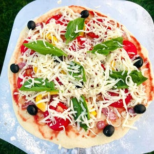 May include: A freshly made pizza on a metal pizza peel, ready for the oven. The pizza is topped with shredded cheese, red tomatoes, yellow peppers, black olives, and fresh basil leaves. The pizza sits on a bed of green grass.