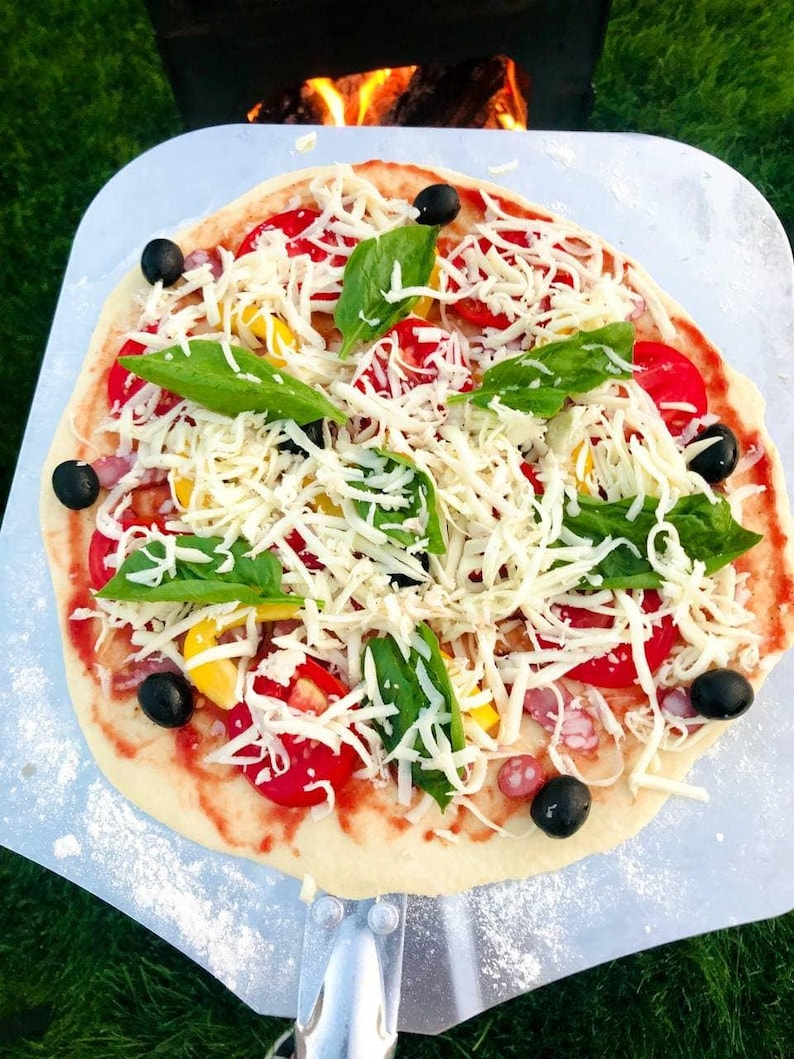 May include: A freshly made pizza on a metal pizza peel, ready for the oven. The pizza is topped with shredded cheese, red tomatoes, yellow peppers, black olives, and fresh basil leaves. The pizza sits on a bed of green grass.