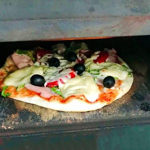 May include: A freshly baked pizza with toppings including black olives, red tomatoes, and melted white cheese. The pizza is on a crust and is inside a dark oven. The pizza is a delicious meal.