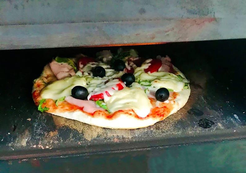 May include: A freshly baked pizza with toppings including black olives, red tomatoes, and melted white cheese. The pizza is on a crust and is inside a dark oven. The pizza is a delicious meal.