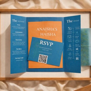 May include: Wedding invitation suite in a wooden frame. The invitation features blue and orange panels with the names "Anaisha" and "Haisha". An RSVP card with a QR code is included. The details and event information are displayed on the panels.
