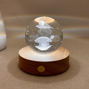 May include: A clear glass sphere with an illuminated seashell and pearl design, set on a wooden base. The sphere is lit from within, creating a soft glow. A small, round, gold-colored button is on the base. A white vase is in the background.