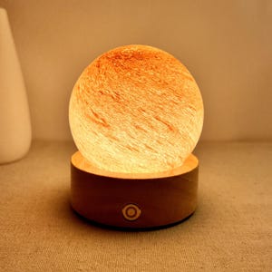 May include: A glowing, spherical lamp with an orange and white marbled glass shade sits atop a round, wooden base. The base has a single, centered button. A white vase is in the background.