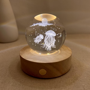 May include: A decorative glass sphere featuring etched jellyfish designs, lit from within. The sphere rests on a round wooden base. The jellyfish are white and the sphere is clear, creating a calming, artistic display.