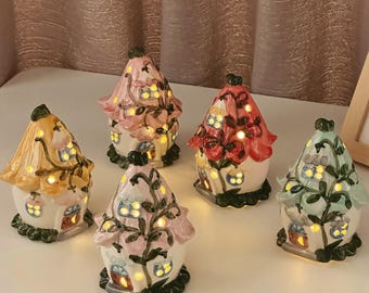 May include: Five ceramic house-shaped lanterns with pointed roofs, each featuring cut-out windows and doors that emit a warm internal glow. The houses are painted in pastel shades of yellow, pink, red, and light green with dark green vine details.