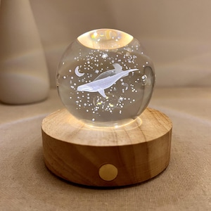 Puede incluir: Una esfera de cristal transparente con un diseño de ballena, luna y estrellas, iluminada por una luz cálida. La esfera se asienta sobre una base de madera redonda. Objeto decorativo.