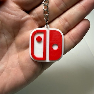 May include: A white and red keychain featuring the design of a video game console controller. The keychain is attached to a silver chain. The controller design has a red outline and two red circles on a white background.