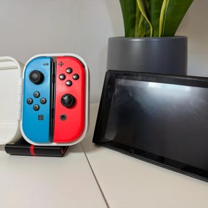 May include: A Nintendo Switch console with blue and red Joy-Con controllers, a black screen, and a white case. A black charging dock is under the controllers. A potted plant is in the background.