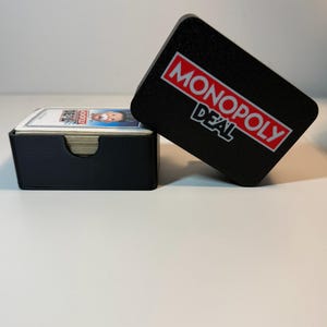 May include: A black card holder and lid with the words "MONOPOLY DEAL" in red and white. The card holder contains a stack of cards. The lid is leaning against the card holder. The game is a card game version of the popular board game.