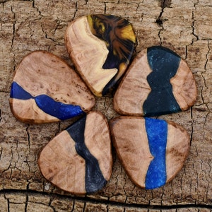 Può includere: Cinque plettri per chitarra in legno con dettagli in resina blu, nera e marrone. I plettri sono disposti in cerchio su una superficie in legno.