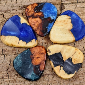 May include: Five guitar picks made of wood and resin with blue, green, and black swirls. The picks are arranged in a circle on a wooden surface.