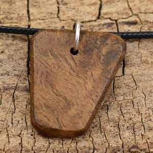 May include: A wooden pendant with a triangular shape and a silver loop. The pendant has a dark brown color and a wood grain pattern. It is hanging from a black cord.