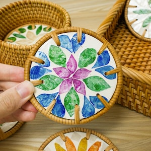 May include: A set of decorative coasters with a floral design. Each coaster features a white ceramic center with a hand-painted flower in shades of blue, green, and purple. The coasters are framed with woven rattan and are displayed in a matching basket.