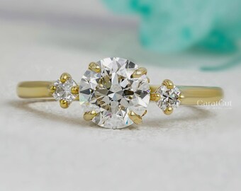 May include: A gold ring featuring a large, round, clear diamond centre stone, with two smaller diamonds either side. The ring is set on a white surface with a blurred background. The text "CaratCut Diamond" is visible.