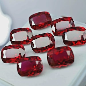 Puede incluir: Una colección de ocho gemas facetadas, de corte cojín, de color rojo intenso. Las gemas están dispuestas sobre una superficie blanca, mostrando sus superficies pulidas y reflejos internos. Las piedras exhiben un color rico y saturado.