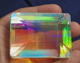May include: A rectangular, iridescent gemstone with a faceted cut. The gem displays a spectrum of colors, including green, yellow, orange, and blue, reflecting light. The stone is held in a hand against a dark blue background.