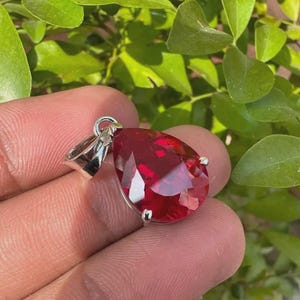 Op de afbeelding: Een peervormige, dieprode gefacetteerde edelsteen hanger in een gepolijste, zilverkleurige metalen zetting. De steen heeft een briljant slijpvorm met meerdere driehoekige facetten die het licht reflecteren voor een levendige uitstraling.