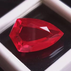 Può includere: Una gemma rossa sfaccettata e vibrante con una forma angolare unica. La gemma è esposta in una scatola di presentazione con cornice bianca e sfondo nero, evidenziando la sua brillantezza e il suo colore. Le sfaccettature catturano la luce.