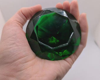 May include: A large, faceted, emerald-green gemstone held in a hand. The gem has a complex, geometric design with multiple facets that reflect light. The gem is a deep, rich green color and appears to be made of glass or a similar material.