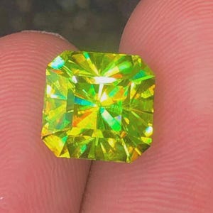 May include: A radiant, square-cut gemstone with a vibrant yellow-green hue. The facets of the gem reflect light, creating a dazzling display of color. The gemstone is held between fingers, showcasing its size and brilliance.