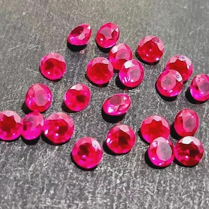 Puede incluir: Una colección de gemas facetadas, de forma ovalada, de color rojo brillante, dispersas sobre una superficie oscura. Las gemas tienen un alto brillo, reflejando la luz y creando un efecto brillante. La disposición sugiere rubíes sueltos.