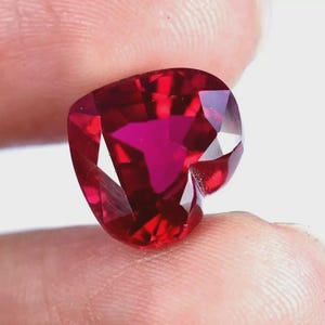 May include: A faceted, heart-shaped gemstone with a deep red hue. The gem's surface reflects light, showcasing its intricate cuts and internal brilliance. The gemstone is held between two fingers.