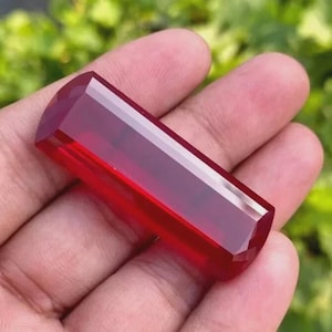 May include: A faceted, rectangular gemstone with a deep red hue. The gem is held in a hand, showcasing its polished surfaces and geometric cuts. The gemstone's color is consistent throughout, with a slight translucence.