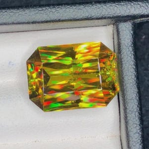 May include: A rectangular-cut gemstone with multiple facets. The gem exhibits a spectrum of colours, including yellow, green, and red, producing a striking visual effect. The stone is presented on a white surface within a black box.