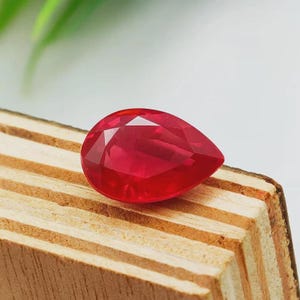 Può includere: Una gemma di rubino sfaccettata a forma di pera. La gemma è di un colore rosso intenso e vibrante e si trova su una superficie di legno con venature visibili. Il taglio della gemma crea molteplici superfici riflettenti.