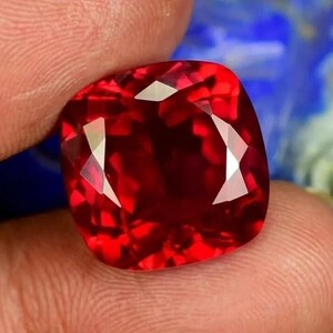 May include: A vibrant, cushion-cut gemstone with a deep red hue. The gem features multiple facets that reflect light, creating a sparkling effect. The stone is held between fingers, showcasing its size and color.