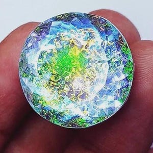 May include: A round, iridescent gemstone with a faceted surface. The gem features a blend of blue, green, and yellow hues, creating a vibrant, multi-colored effect. The gemstone is held in a hand, showcasing its size and brilliance.