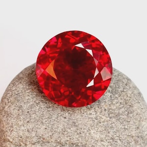 May include: A round, faceted gemstone with a vibrant, deep red colour. The stone features a brilliant cut with numerous triangular and kite-shaped facets that reflect light, creating a complex pattern of bright red highlights and dark shadows.