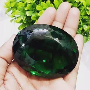 May include: A large, faceted, oval-shaped gemstone in a deep emerald green hue. The gem is held in an open hand, with a backdrop of green foliage. The cut of the stone creates multiple reflective surfaces.