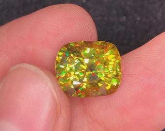 May include: A cushion-cut gemstone with a vibrant yellow hue, displaying internal flashes of red, green, and orange. The gem is held between fingers, showcasing its brilliance and clarity. The stone's facets catch the light, creating a dazzling effect.