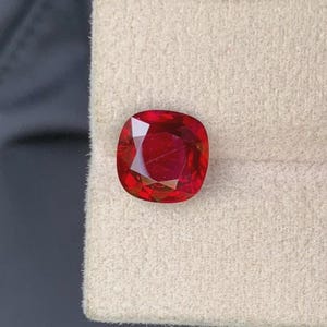 May include: A cushion-cut ruby gemstone, displaying a rich, deep red hue. The facets of the stone reflect light, creating a sparkling effect. The gem is presented against a neutral backdrop, emphasizing its colour and clarity. A fine example of a precious stone.