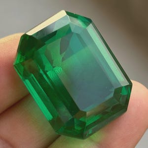 May include: A large, emerald-cut gemstone with a deep green hue. The gem has a rectangular shape with beveled edges, reflecting light and creating a brilliant sparkle. The stone is held in a hand, showcasing its size and clarity.