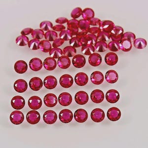 Puede incluir: Una colección de numerosas piedras preciosas pequeñas, redondas y facetadas en un color rojo rosado intenso y vibrante. Las piedras están dispuestas en un montón suelto en la parte superior y organizadas en filas horizontales de siete abajo.