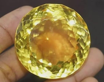 May include: A faceted, round, yellow gemstone with a warm, golden center. The gem is held in a hand, showcasing its brilliant cut and clarity. The stone's color ranges from light yellow to a deeper amber hue.