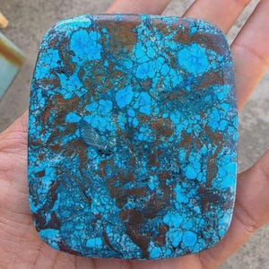 Pode incluir: Uma pedra retangular polida com um padrão vibrante de azul turquesa e castanho. A superfície da pedra é um mosaico de cores, com o azul a dominar e o castanho a criar veios intrincados. As bordas são arredondadas, dando-lhe um aspeto liso.