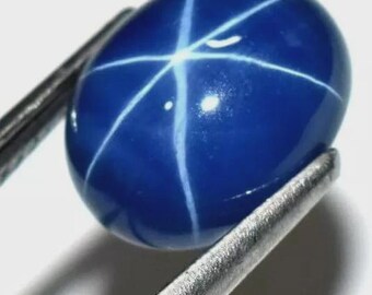 May include: A polished, oval-shaped, deep blue gemstone with a six-rayed star pattern. The star is formed by white, intersecting lines across the surface of the gem. The stone is held by a pair of metal tweezers, showcasing its brilliance.