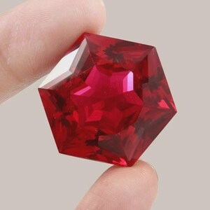 May include: A faceted, pentagonal-shaped gemstone with a deep, vibrant red color. The stone features multiple triangular and trapezoidal facets that create a complex, geometric pattern across its surface, reflecting light with high clarity and brilliance.