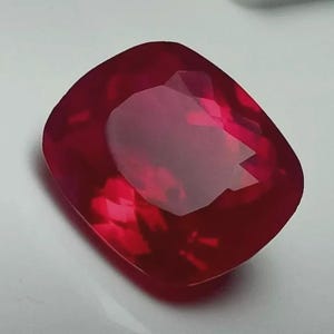 Peut inclure: Une pierre précieuse taillée en coussin, de couleur rouge profond. La gemme a une surface polie et un intérieur translucide de couleur plus claire. La forme est un carré modifié aux coins arrondis, reflétant la lumière.