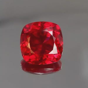 Puede incluir: Una gema roja de talla cojín con una superficie facetada que refleja la luz. La piedra muestra un tono carmesí profundo y saturado en toda su estructura, exhibiendo cortes geométricos precisos que crean múltiples reflejos internos.