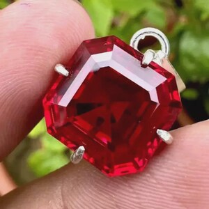 Peut inclure: Un pendentif en pierre précieuse rouge foncé, facetté et octogonal. La gemme est sertie dans un sertissage en métal argenté avec une petite boucle en haut pour une chaîne. Le pendentif est tenu entre les doigts sur un fond vert flou.